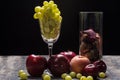 Still life of a glass with grapes, apple, Peach, Plumbs, and a glass of potpourri sitting on wooden table with black background.