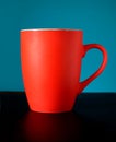 Still life with high red porcelain tea cup or coffee cup indoors studio shot on black surface over cyan background front view clos