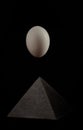 Still-life. A large white egg hangs above a black pyramid on a black background with a reflection