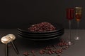 Still life of a stack of black plates with red beans, spoons and glasses of colored glass on a black background Royalty Free Stock Photo