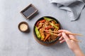 Stir-fry with soba noodles, meat and vegetables in black plate on a light background with hand hold chopsticks