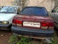 Stock photo of abandoned broken , damaged maroon color car left in the scrap yard for recycling . White color and gray color car