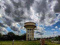 Old rusty yellow painted water tank tower surrounded by green trees under blue sky with white clouds at the middle of the land. Royalty Free Stock Photo