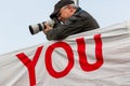 Senior male taking photographs using a long lens. Leaning on a large sign with the word You.