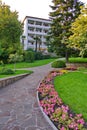 A stone path with planted flowers, tall green trees and a lawn leads to a white five-story building with balconies