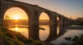 Stone railway viaduct with multiple arches spans a tranquil river. The sun sets in the background Royalty Free Stock Photo