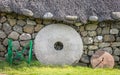 A stone wall with a thatched roof. a large stone millstone leaning on a wall