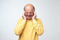 Mature man in yellow tshirt suffering from loud music and pain screaming trying to plug ears against grey background. Royalty Free Stock Photo