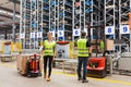 Storehouse employees in uniform standing near pallet truck and forklift in modern automatic warehouse.Boxes are on the shelves of Royalty Free Stock Photo