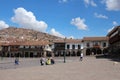 Stores lining the Plaza de Armas with tourists walking and sitting, with a hillside filled with homes in Cusco, Peru