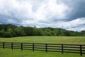 Storm clouds over a brilliant green field, black rail fence in front with treeline on the horizon Royalty Free Stock Photo