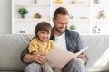 Storytime. Young loving father reading fairy tales to his cute little son, sitting with big child book on sofa at home