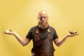 Comic portrait of muscular bearded bald man, blacksmith in leather apron or uniform isolated on yellow studio background