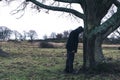 A strange surreal picture of a mysterious hooded man learning against a tree on a bleak moody winters day in the countryside