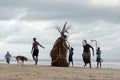 A straw scarecrow stands on a sandy beach on the shore of the ocean among people, during a ceremony for good weather and good