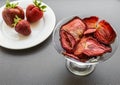 Strawberry chips in a glass vase on a leg next to three fresh strawberries on a white glass plate on a gray background Royalty Free Stock Photo