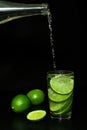 Stream of cold water from a bottle is poured into a glass with ice and fresh ripe slice green limes on black background.