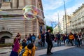 Street artist dressed like British gentleman in black costume with hat cylinder performing for the kids blowing huge soap bubbles