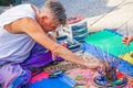 Street artist in t shirt and bright pants sit on carpet emptying little bottle of blue paint into saucer with brushes in mug