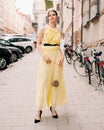 Street fashion. Beautiful young model lady walking on city road. Fashionable feminine look. Long yellow summer evening dress. Royalty Free Stock Photo