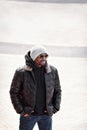 Street fashion, portrait stylish man in black leather jacket, hoodie, knitted hat and sunglasses standing, hands in Royalty Free Stock Photo