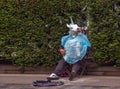 A street musician in a unicorn mask sits on the sidewalk and plays the accordion asking for money