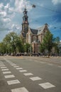 Street with old brick church steeple, trees and people passing by under cloudy blue sky in Amsterdam. Royalty Free Stock Photo