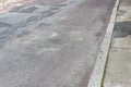 Street and sidewalk in crumbling disrepair, heavily patched asphalt roadway