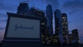 Street signage board with Johnson`s logo in the evening. Blurred business district skyscrapers background. Editorial 3D