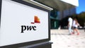 Street signage board with PricewaterhouseCoopers PwC logo. Blurred office center and walking people background
