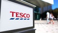 Street signage board with Tesco logo. Blurred office center and walking people background. Editorial 3D rendering