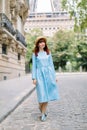 Street view of pretty young red haired lady, wearing elegant blue dress and straw hat, walking during the morning in Royalty Free Stock Photo