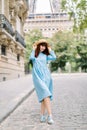Street view of pretty young red haired lady, wearing elegant blue dress and straw hat, walking during the morning in Royalty Free Stock Photo