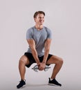 Strength workout concept. Positive young sportsman with dumbbell doing squat on light studio background, full length Royalty Free Stock Photo