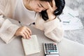 Stress Asian woman calculating her debt having problem with loan looking at list of expenses on notepad with invoices on floor