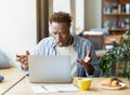 Stressed African American guy in headphones using laptop to work on complicated business project, facing problem at cafe