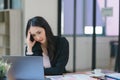 The stressed and exhausted millennial Asian businesswoman is seen sitting at her office desk with her hand on her head