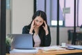 The stressed and exhausted millennial Asian businesswoman is seen sitting at her office desk with her hand on her head