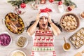 Stressed female cook in a Christmas apron and Santa hat holding her head and lying on the floor surrounded by gingerbread, Linz