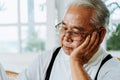 Stressed senior Asian male in glasses leaning on hand and pondering about problems at home. Elderly man thinking about Royalty Free Stock Photo