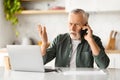 Stressed Senior Man Talking On Cellphone And Using Laptop In Kitchen Royalty Free Stock Photo