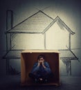 Stressed teenage boy sitting inside a cardboard box hut, imagine a big house. Improvised shelter of homeless and refugees. Poverty