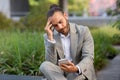 Stressed young businessman looking at smartphone screen while sitting outdoors Royalty Free Stock Photo