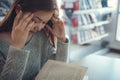 Stressed young female student sitting at wooden table and reading book during exam preparation in library Royalty Free Stock Photo