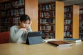 Stressed young female student sitting at wooden table and reading book during exam preparation in library Royalty Free Stock Photo