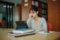 Stressed young female student sitting at wooden table and reading book during exam preparation in library Royalty Free Stock Photo