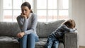 Stressed young mommy sitting separate on couch with offended son.