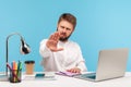 Strict bossy businessman with beard sitting in office workplace with laptop showing stop, warning with hand gesture, access denied