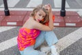 Striking blonde in blouse in coral orange tones with bare shoulder, jeans and high white shoes is sitting on road .