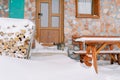 Striped cat stands on the snow-covered threshold of a stone house and looks at a woodpile of firewood